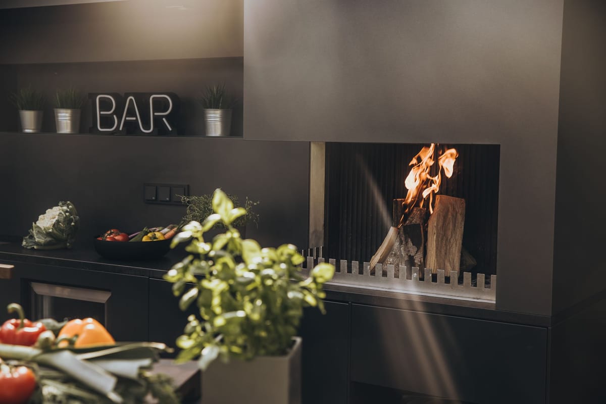 Dark gray outdoor kitchen with an open wood fire in a steel firebox, herbs and vegetables on the counter in soft light
