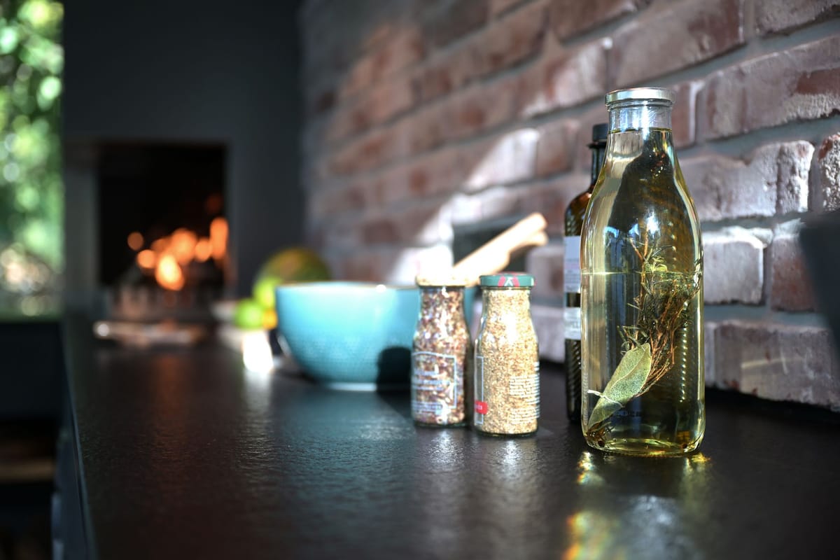 Spice jars and a bottle of herb-infused oil on a black countertop by a brick wall, fire glowing in the background