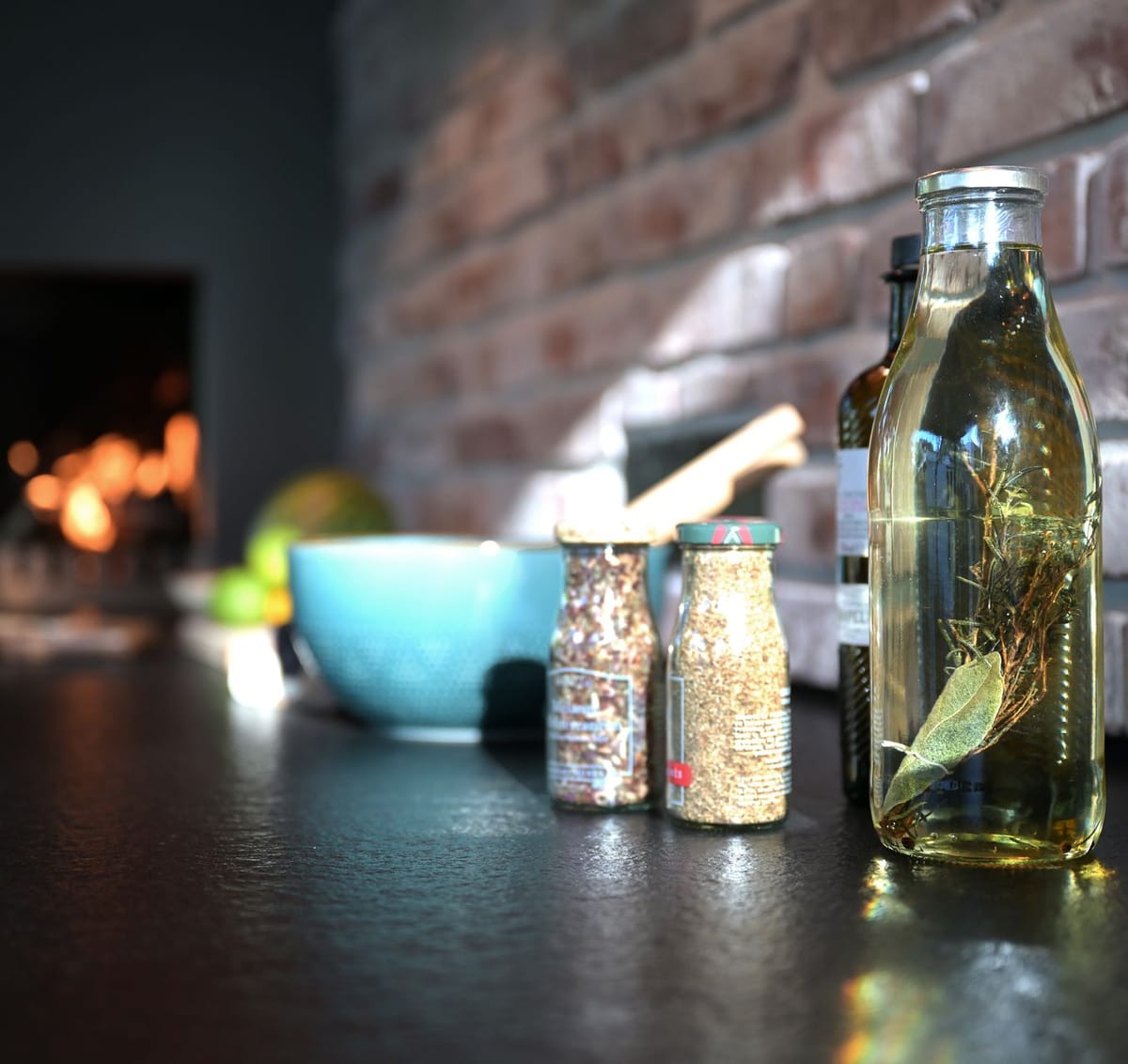 Spice jars and a bottle of herb-infused oil on a black countertop by a brick wall, fire glowing in the background