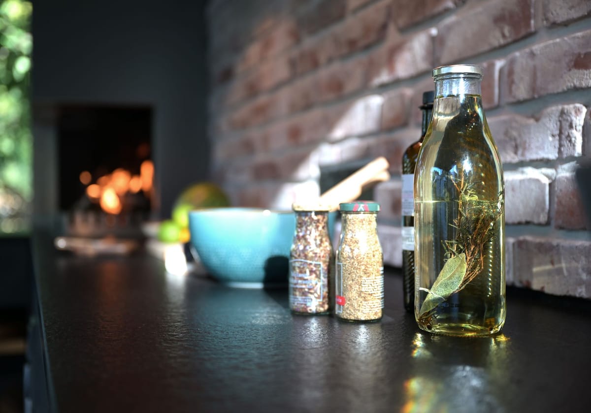 Spice jars and a bottle of herb-infused oil on a black countertop by a brick wall, fire glowing in the background