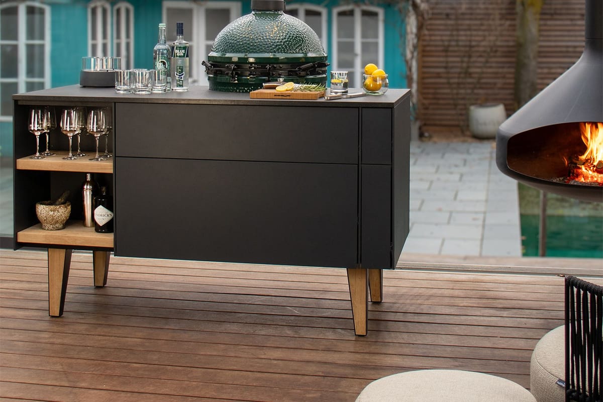 Back of an outdoor island with open oak shelves for glasses and bottles, Big Green Egg set into the worktop, poufs on a wood deck