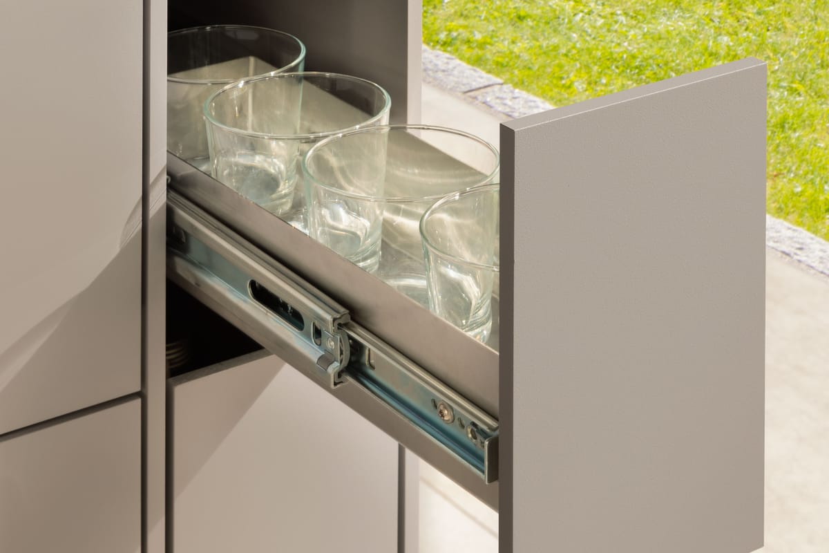 Narrow pull-out drawer with drinking glasses on full-extension runners, taupe front and black stone countertop