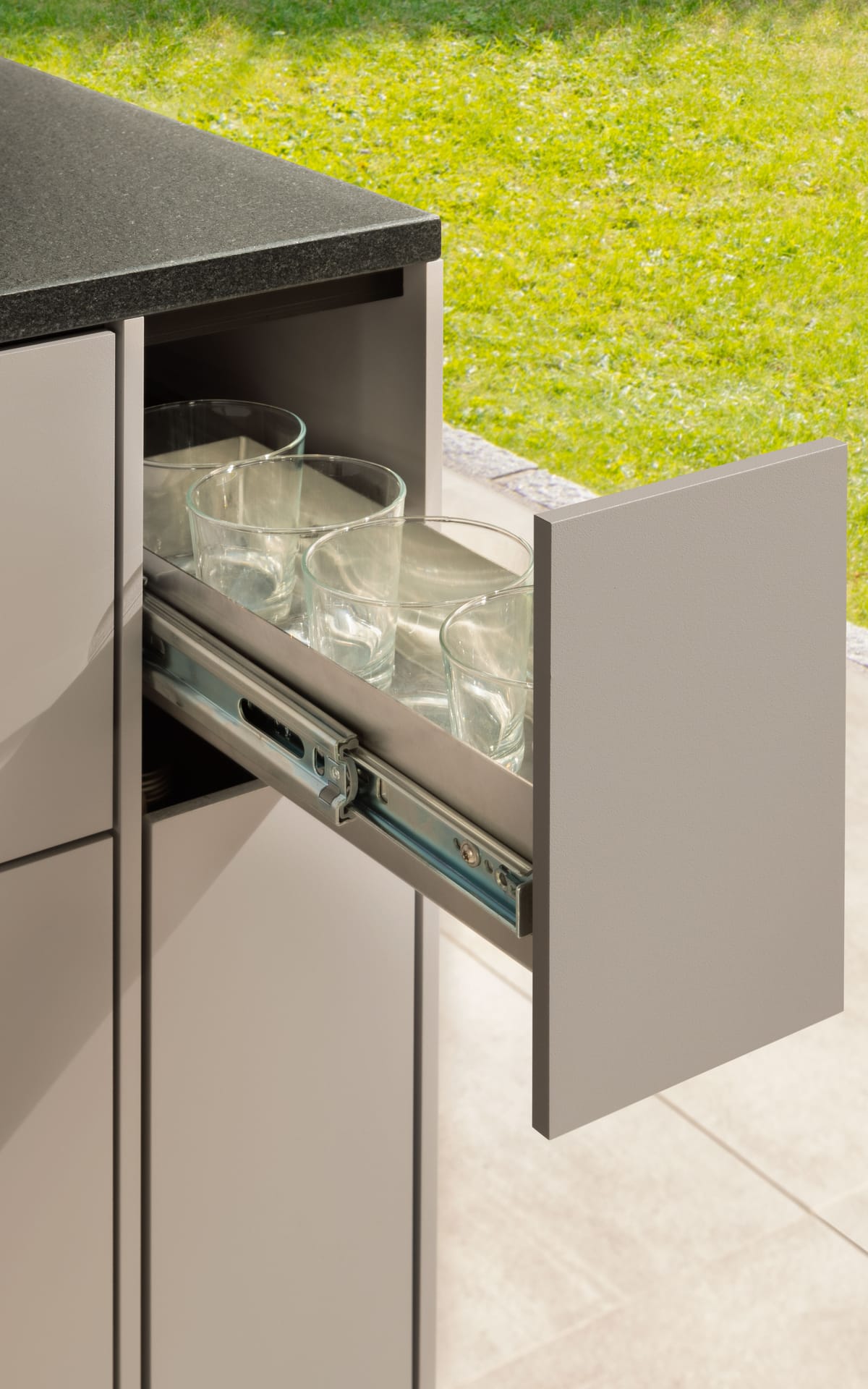 Narrow pull-out drawer with drinking glasses on full-extension runners, taupe front and black stone countertop