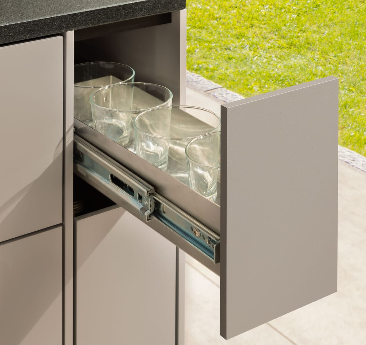 Narrow pull-out drawer with drinking glasses on full-extension runners, taupe front and black stone countertop