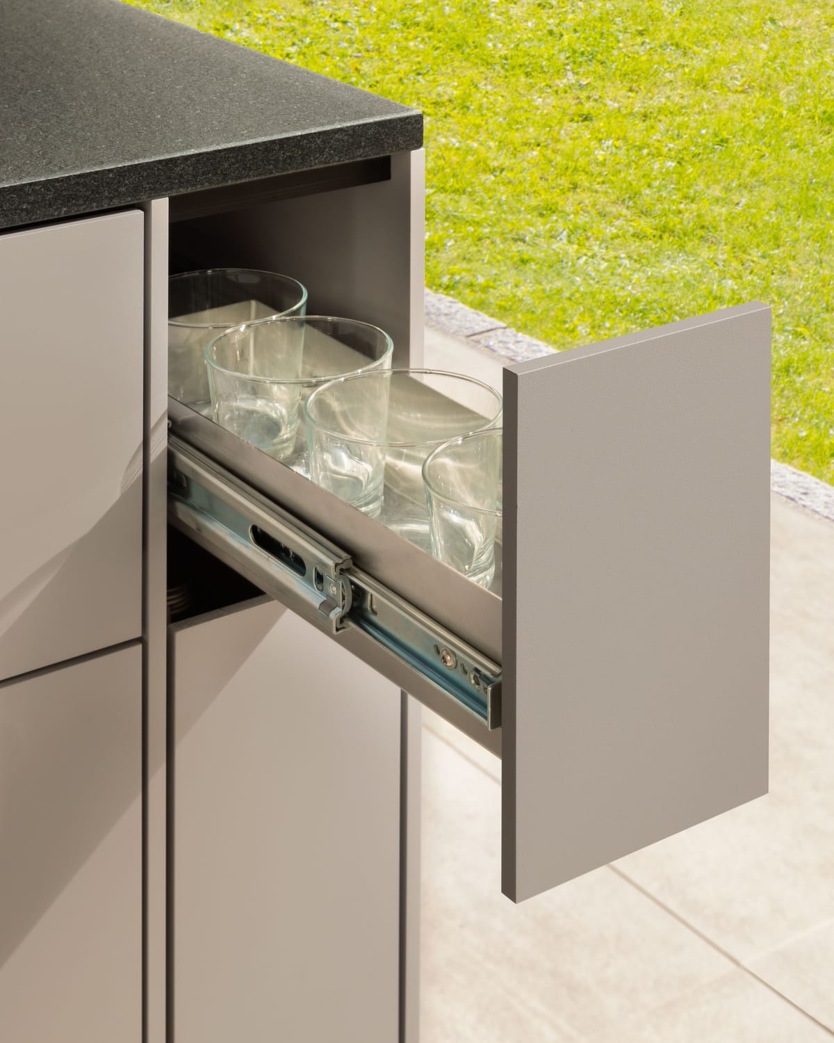 Narrow pull-out drawer with drinking glasses on full-extension runners, taupe front and black stone countertop