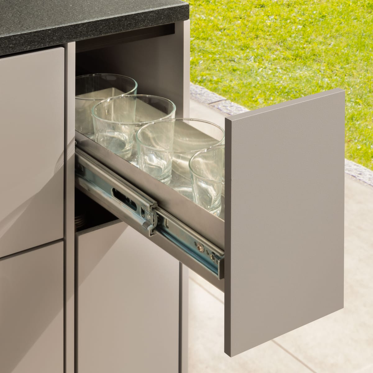 Narrow pull-out drawer with drinking glasses on full-extension runners, taupe front and black stone countertop