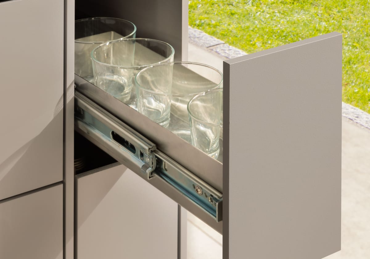 Narrow pull-out drawer with drinking glasses on full-extension runners, taupe front and black stone countertop