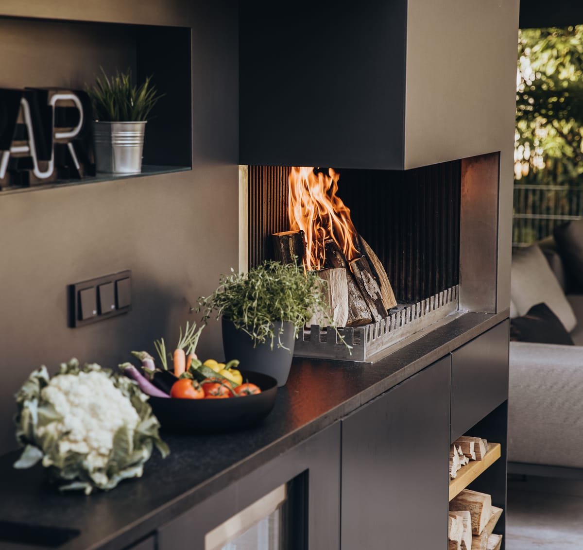 Wood fire in the grill-and-fire element beside herbs and a vegetable bowl on a dark stone counter