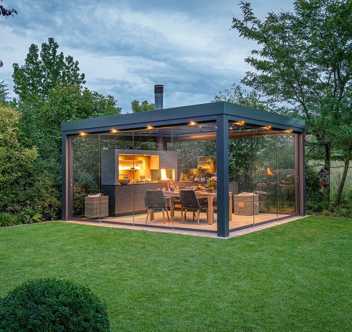 Glass-walled pergola in a garden at dusk with a lit outdoor kitchen, wood fire and dining table inside