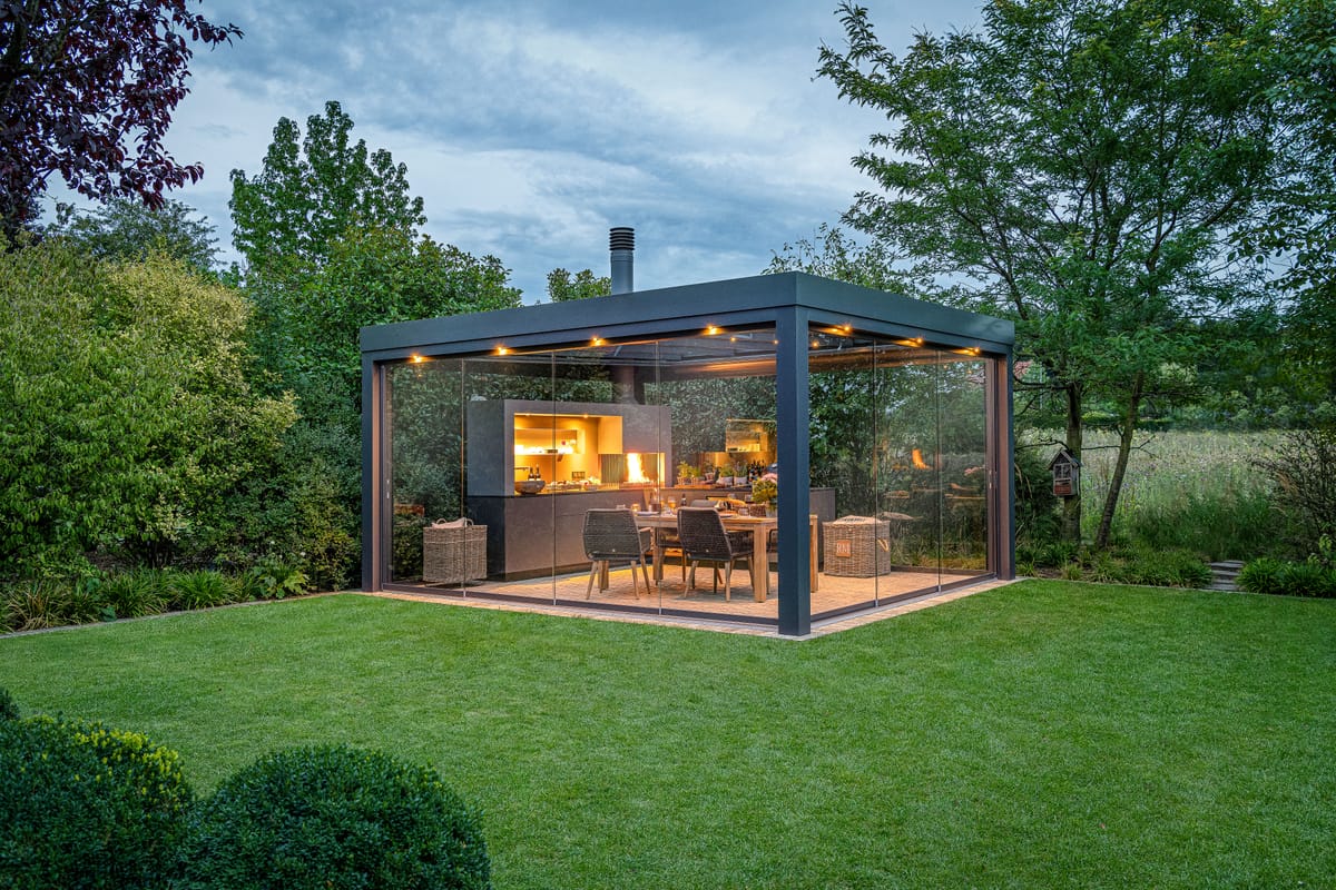 Glass-walled pergola in a garden at dusk with a lit outdoor kitchen, wood fire and dining table inside