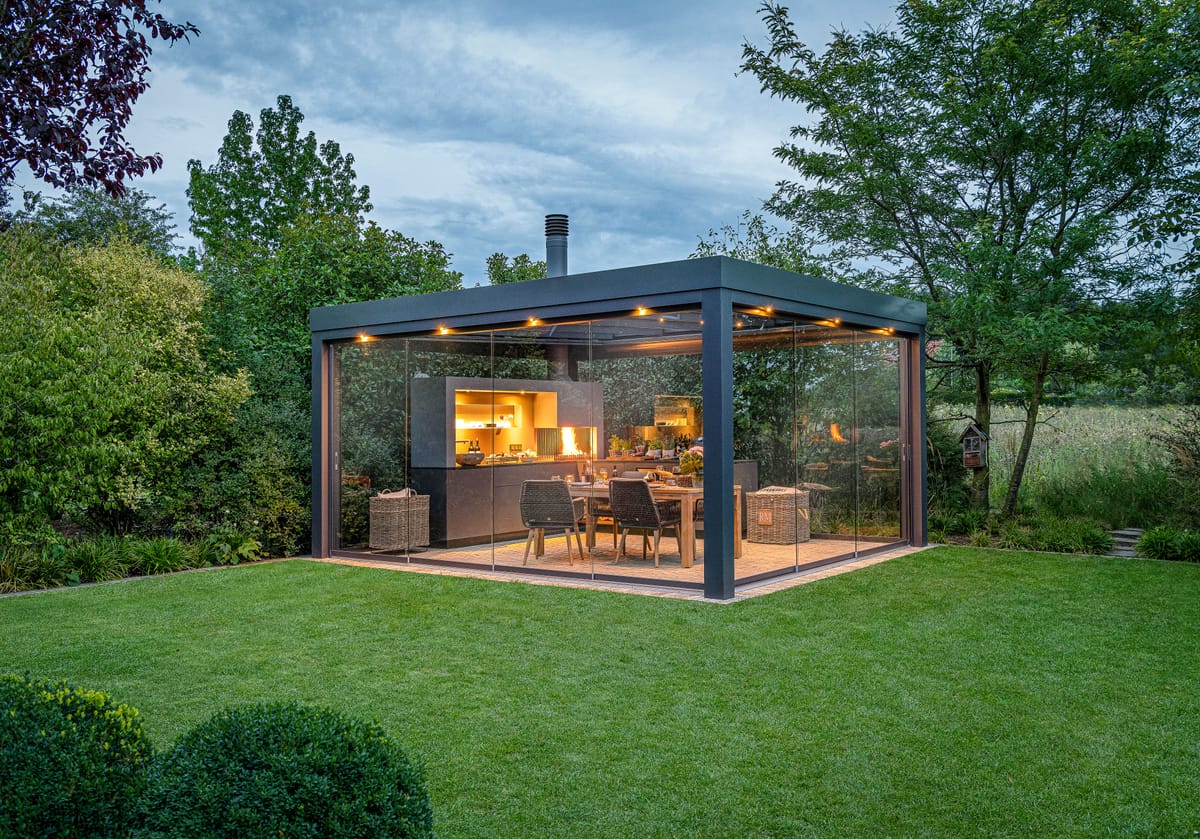 Glass-walled pergola in a garden at dusk with a lit outdoor kitchen, wood fire and dining table inside