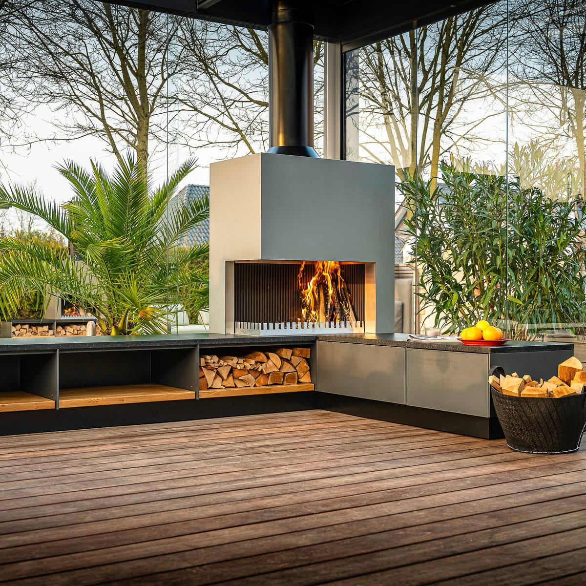 Gray outdoor kitchen with a wood fire in the grill-and-fire element, firewood bench and a potted palm on a deck