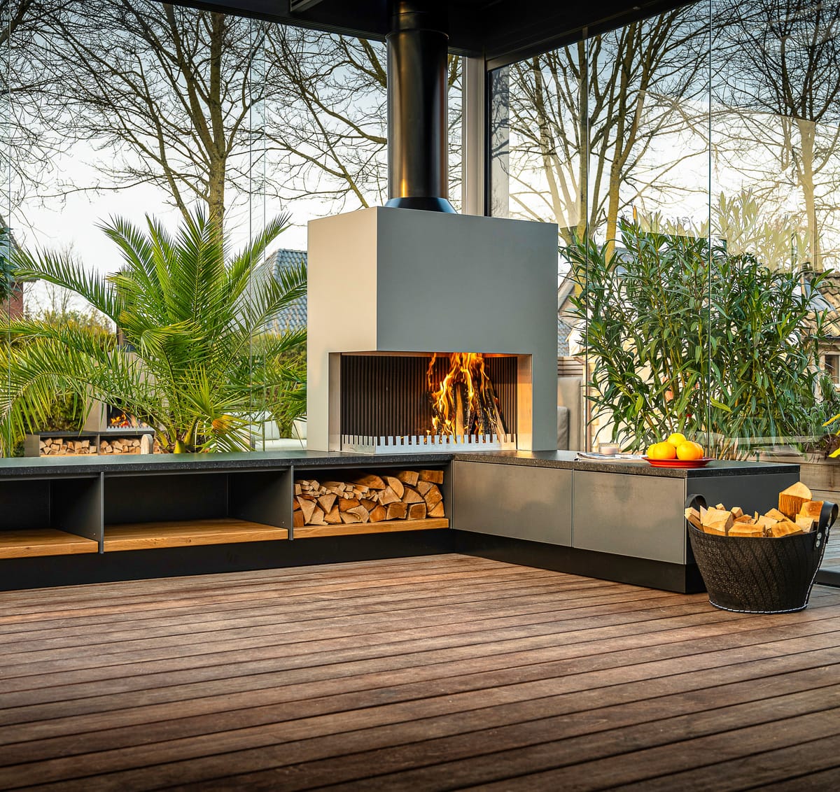 Gray outdoor kitchen with a wood fire in the grill-and-fire element, firewood bench and a potted palm on a deck