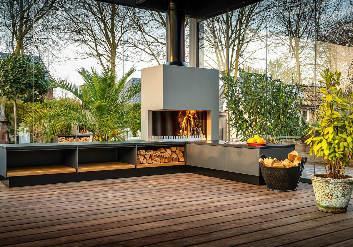 Gray outdoor kitchen with a wood fire in the grill-and-fire element, firewood bench and a potted palm on a deck