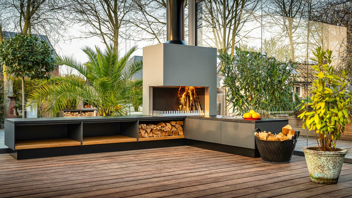Gray outdoor kitchen with a wood fire in the grill-and-fire element, firewood bench and a potted palm on a deck