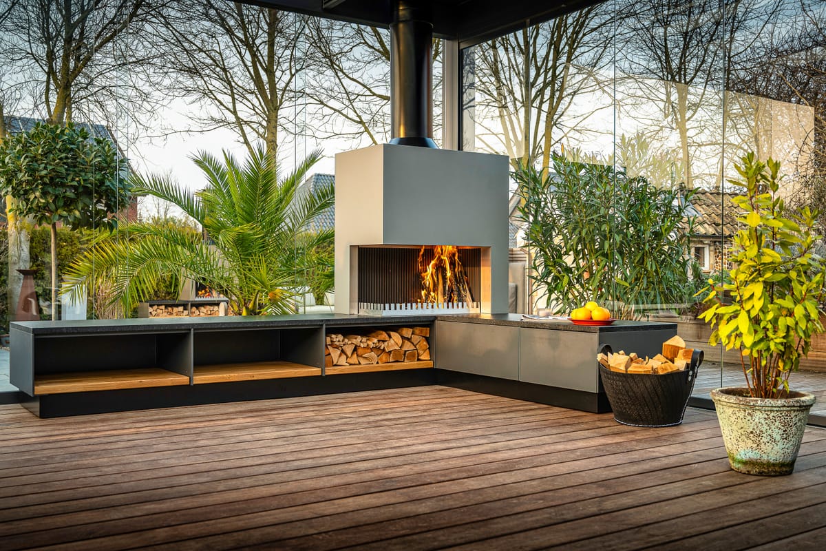 Gray outdoor kitchen with a wood fire in the grill-and-fire element, firewood bench and a potted palm on a deck