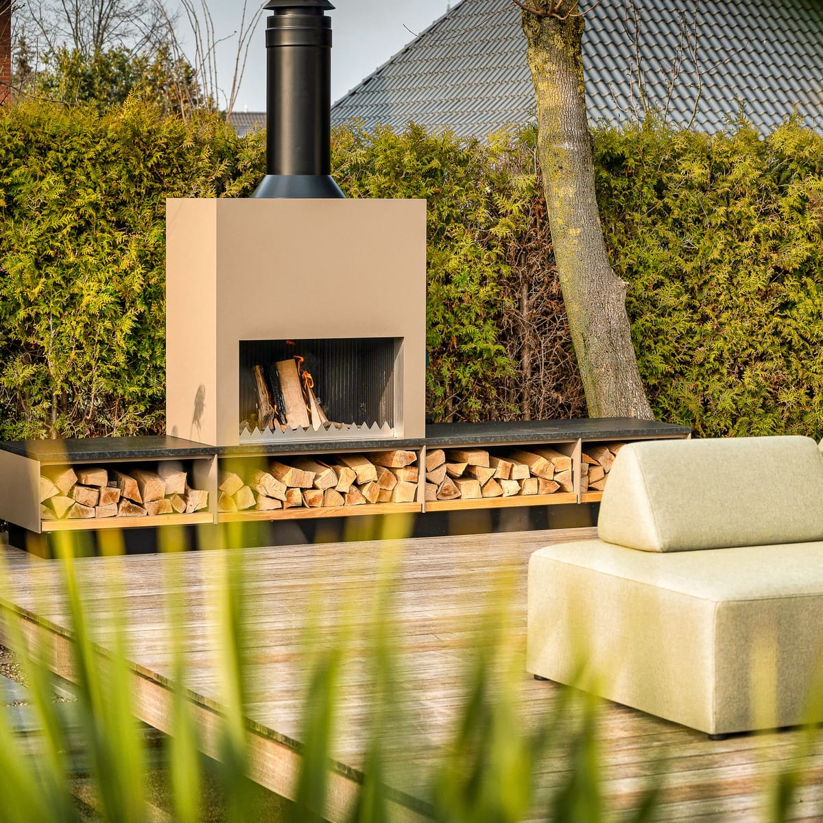 Beige outdoor fireplace with a firewood storage bench on a wooden deck, green lounge sofa in front of a hedge