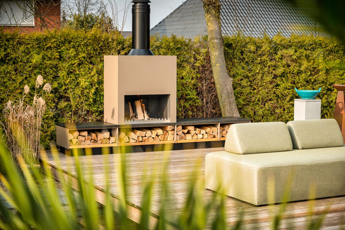 Beige outdoor fireplace with a firewood storage bench on a wooden deck, green lounge sofa in front of a hedge