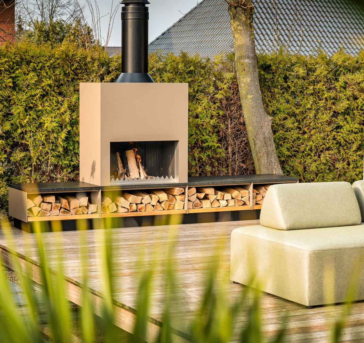 Beige outdoor fireplace with a firewood storage bench on a wooden deck, green lounge sofa in front of a hedge