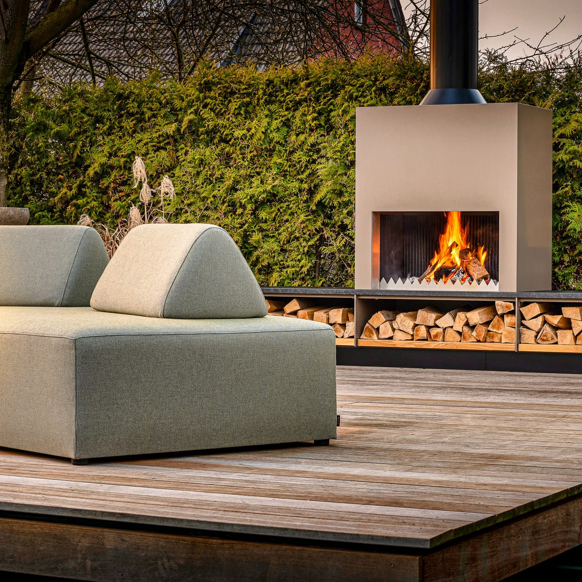 Outdoor fireplace with a burning wood fire and firewood bench behind a green lounge sofa on a wooden deck