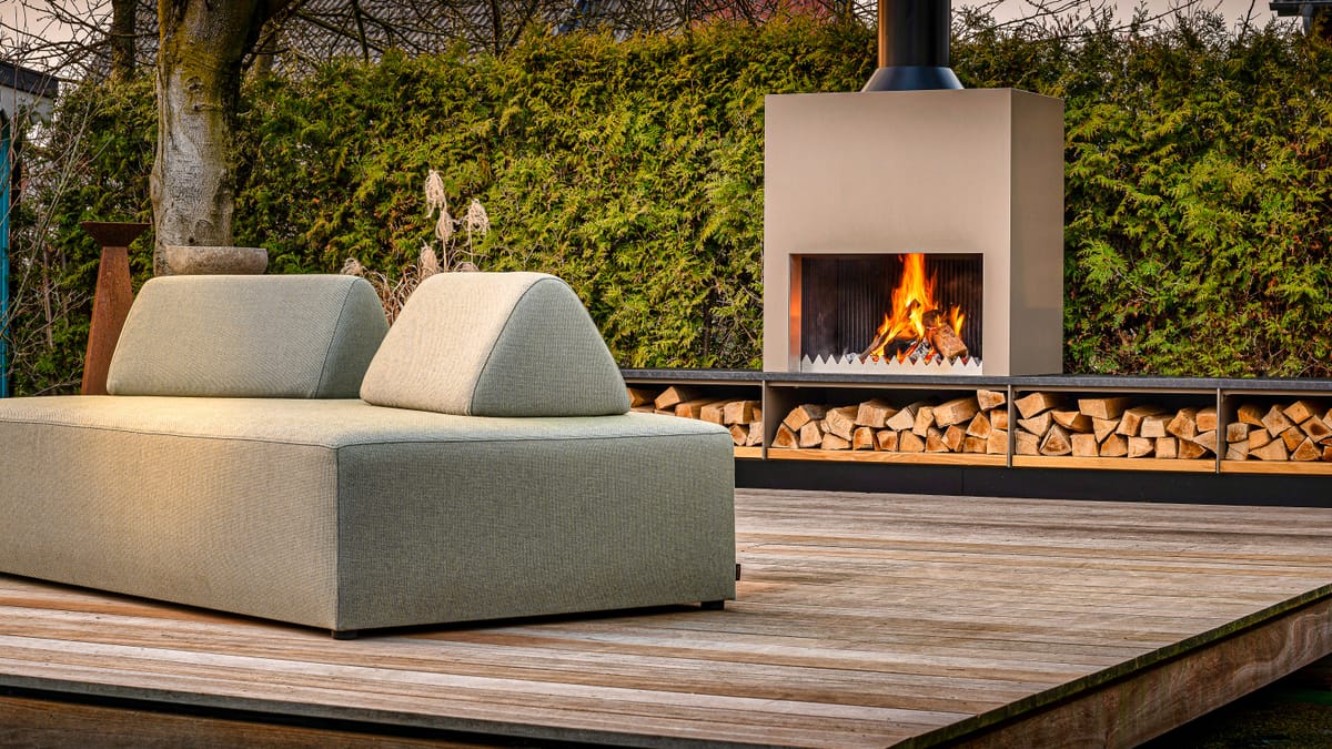Outdoor fireplace with a burning wood fire and firewood bench behind a green lounge sofa on a wooden deck
