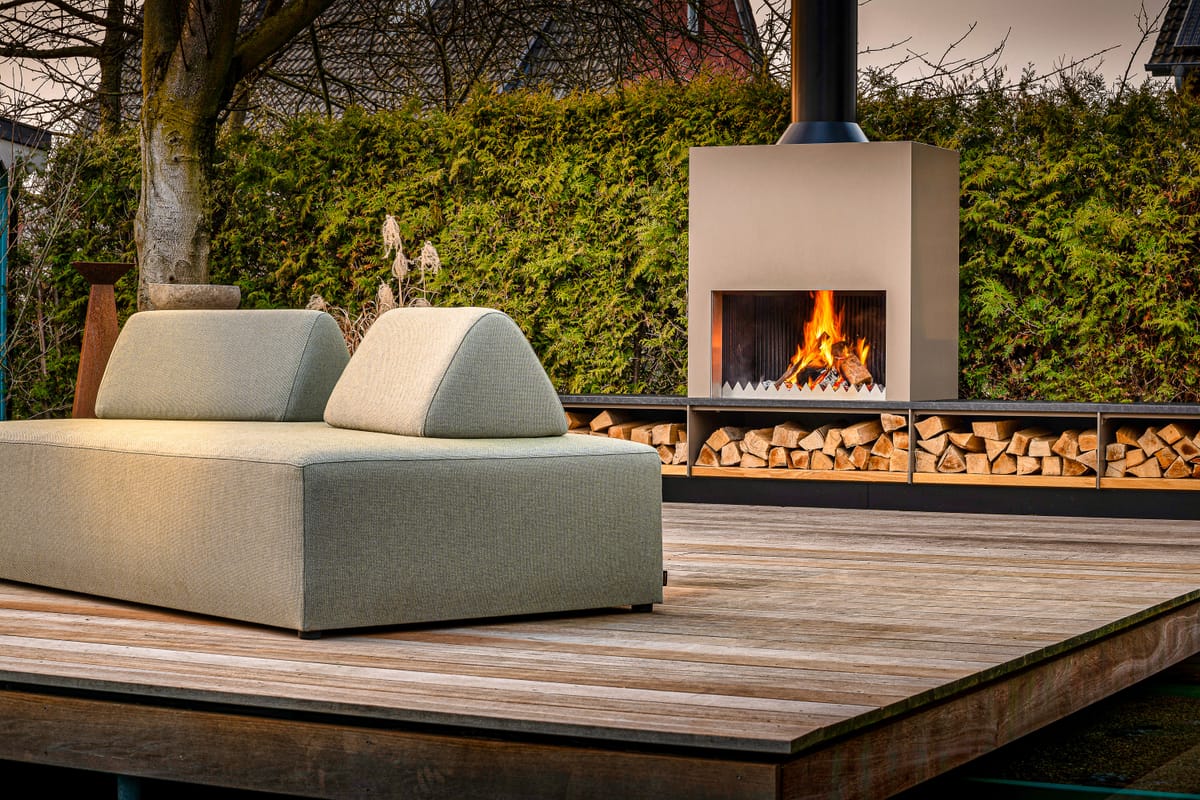 Outdoor fireplace with a burning wood fire and firewood bench behind a green lounge sofa on a wooden deck