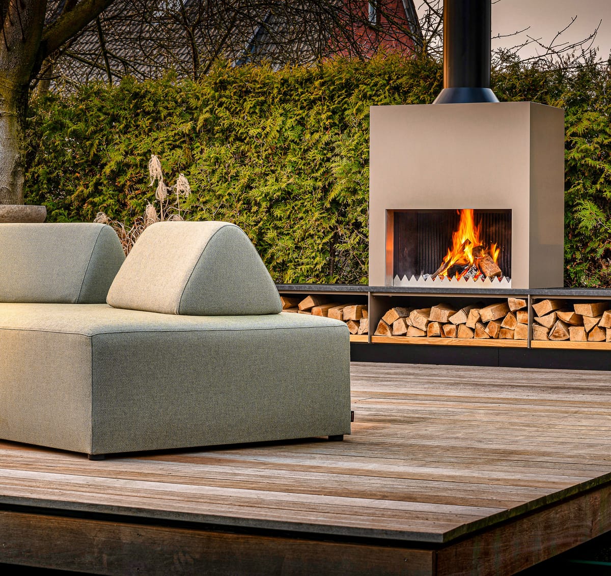 Outdoor fireplace with a burning wood fire and firewood bench behind a green lounge sofa on a wooden deck