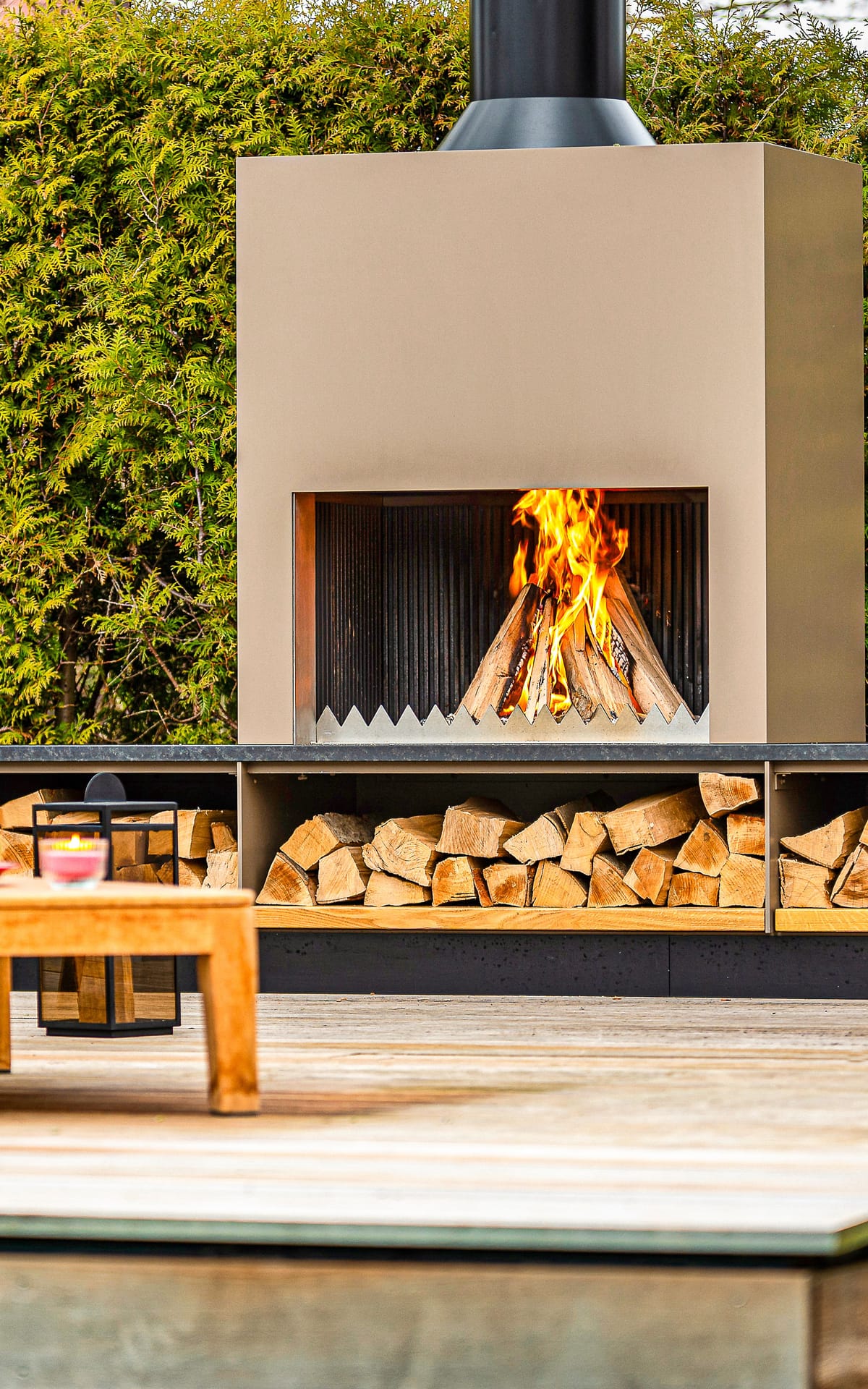 Outdoor fireplace with a wood fire, stacked firewood and blue lounge seating with a teak table on a deck