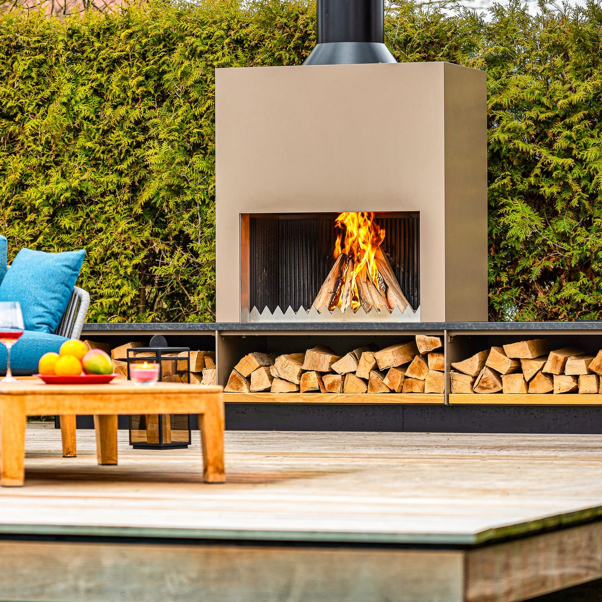 Outdoor fireplace with a wood fire, stacked firewood and blue lounge seating with a teak table on a deck