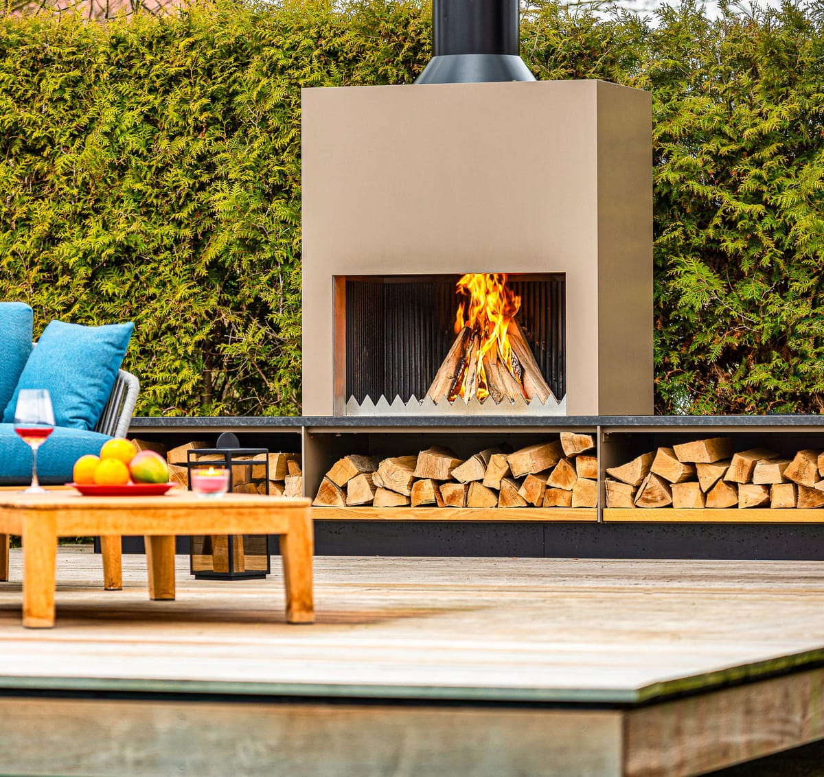 Outdoor fireplace with a wood fire, stacked firewood and blue lounge seating with a teak table on a deck