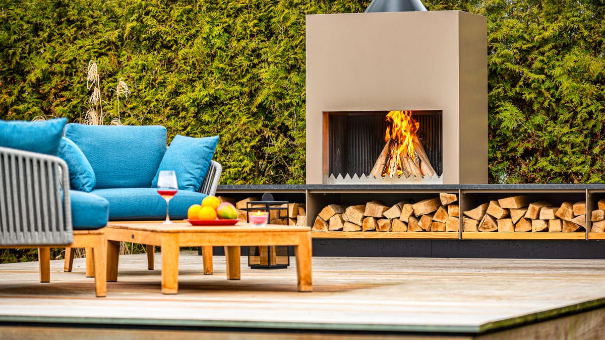 Outdoor fireplace with a wood fire, stacked firewood and blue lounge seating with a teak table on a deck