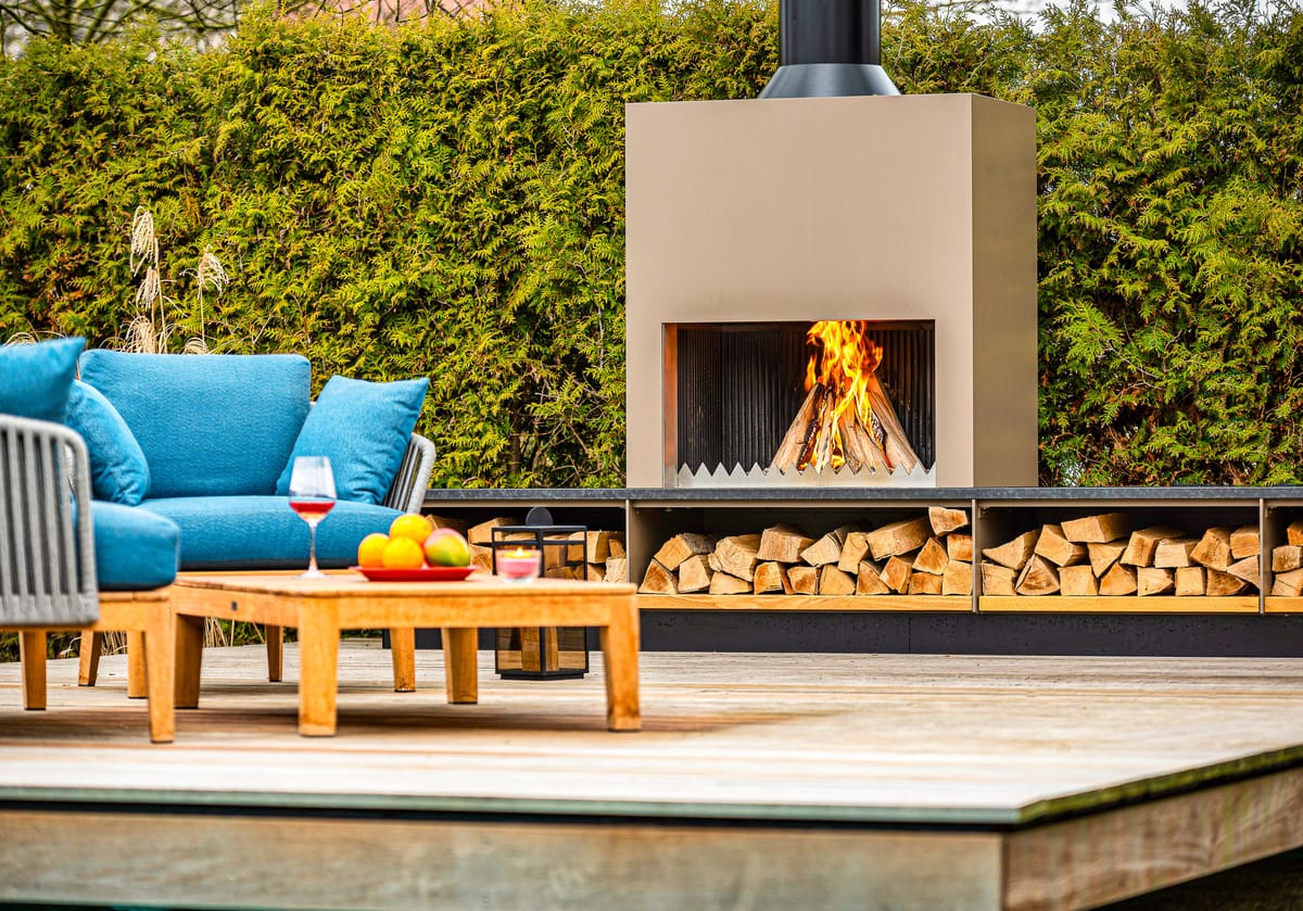 Outdoor fireplace with a wood fire, stacked firewood and blue lounge seating with a teak table on a deck