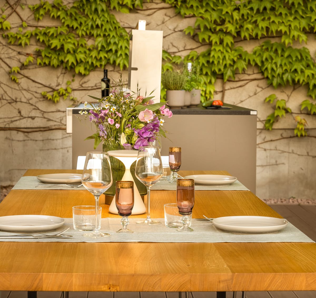 Set dining table with flowers and glasses in front of a taupe outdoor kitchen island by an ivy-covered wall