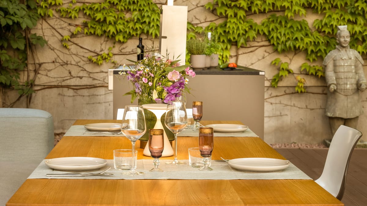 Set dining table with flowers and glasses in front of a taupe outdoor kitchen island by an ivy-covered wall