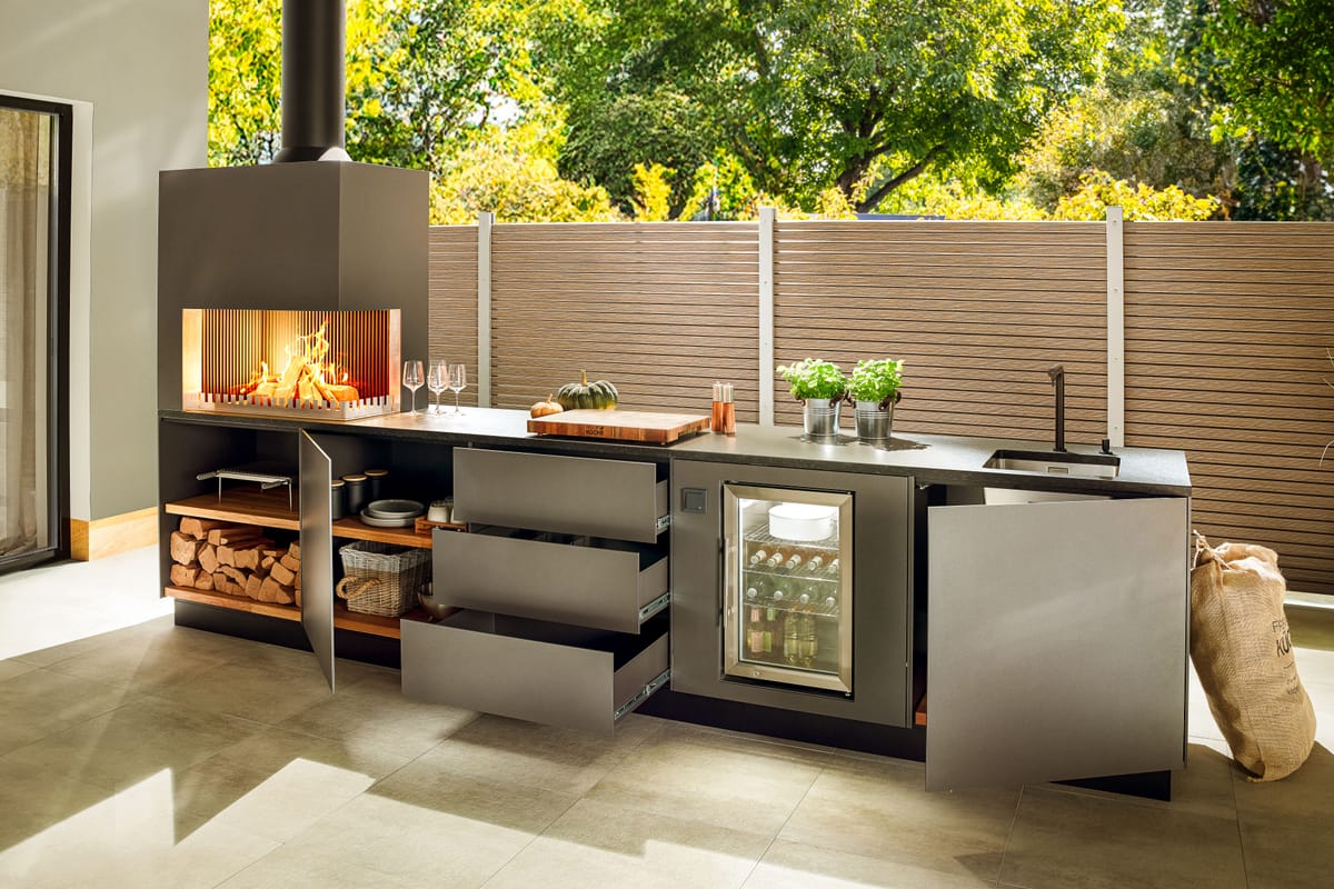 Dark gray outdoor kitchen with open drawers and doors, firewood storage, wood fire and glass-door cooler