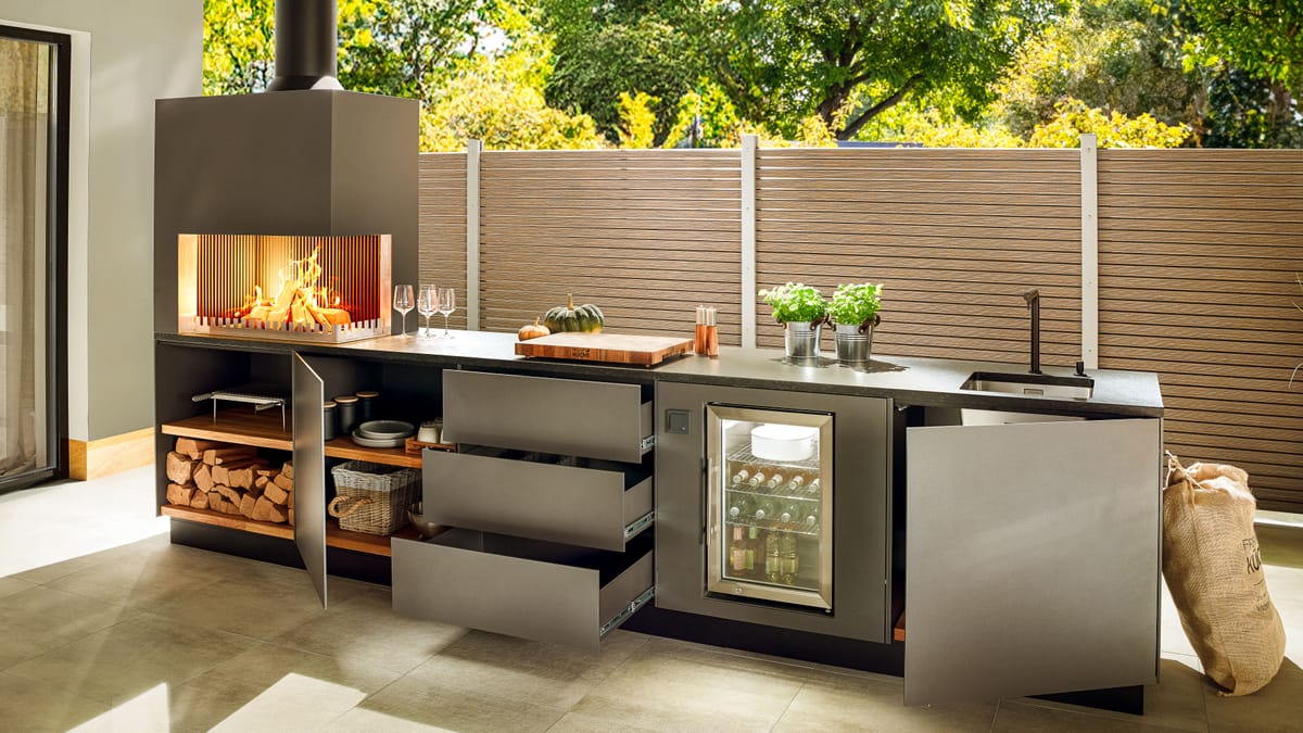 Dark gray outdoor kitchen with open drawers and doors, firewood storage, wood fire and glass-door cooler
