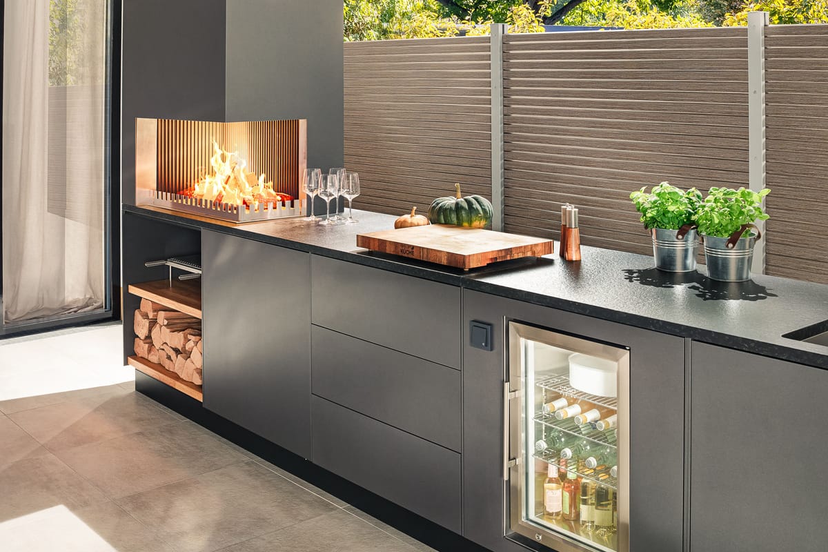Dark gray outdoor kitchen with a wood fire, cutting board, herbs and a glass-door cooler in low sunlight