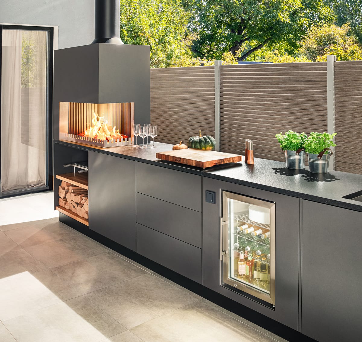 Dark gray outdoor kitchen with a wood fire, cutting board, herbs and a glass-door cooler in low sunlight