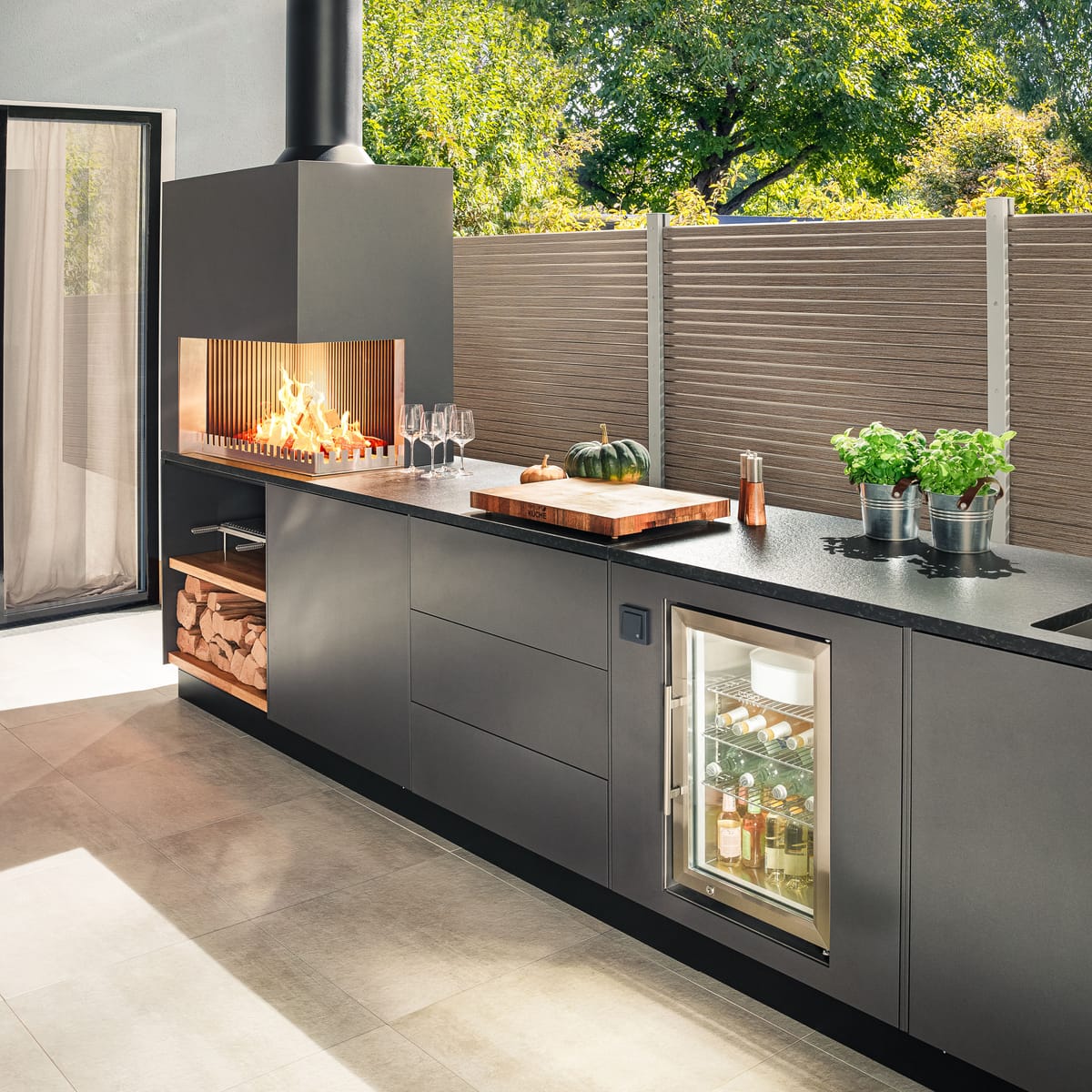 Dark gray outdoor kitchen with a wood fire, cutting board, herbs and a glass-door cooler in low sunlight