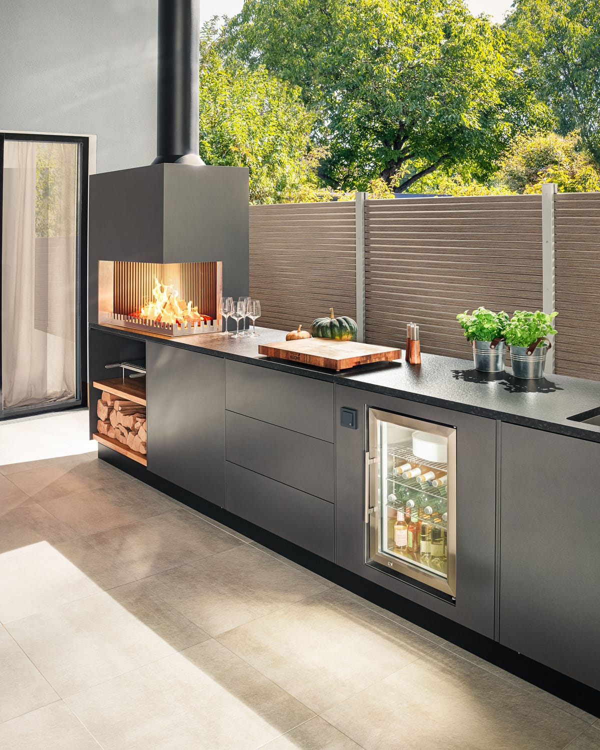 Dark gray outdoor kitchen with a wood fire, cutting board, herbs and a glass-door cooler in low sunlight