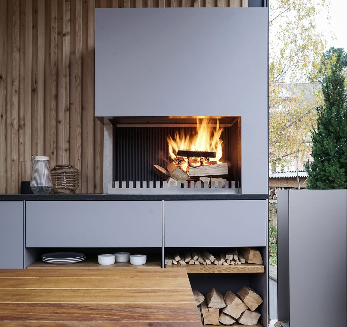 Wood fire in a light gray grill-and-fire element above open shelves with plates and firewood