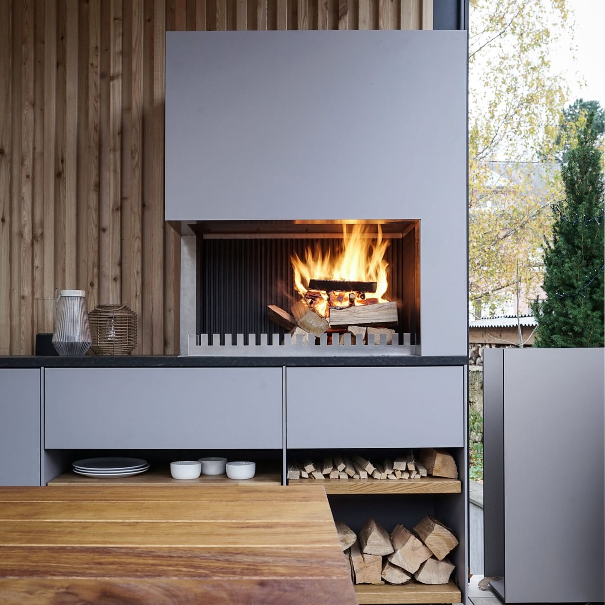 Wood fire in a light gray grill-and-fire element above open shelves with plates and firewood