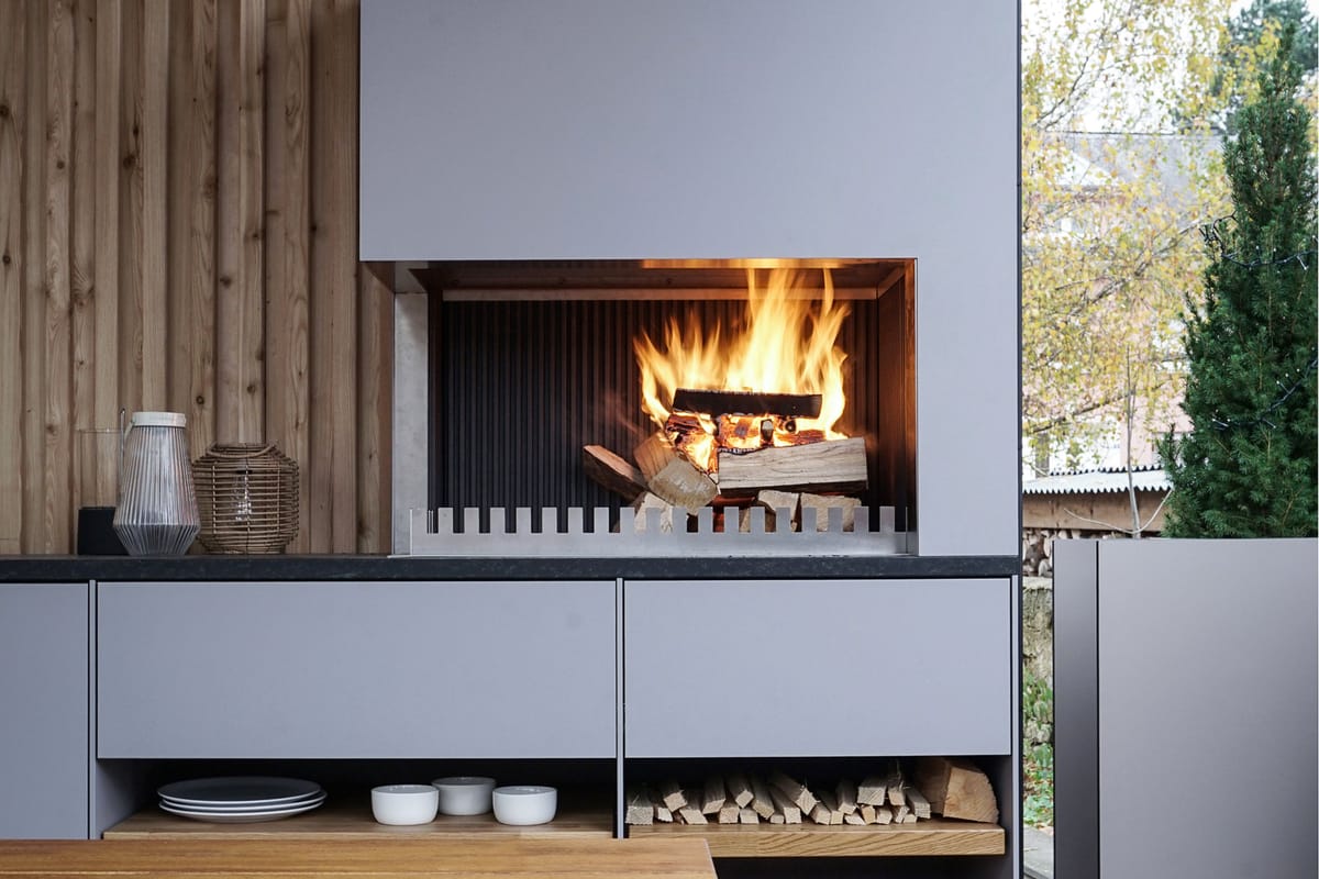 Wood fire in a light gray grill-and-fire element above open shelves with plates and firewood