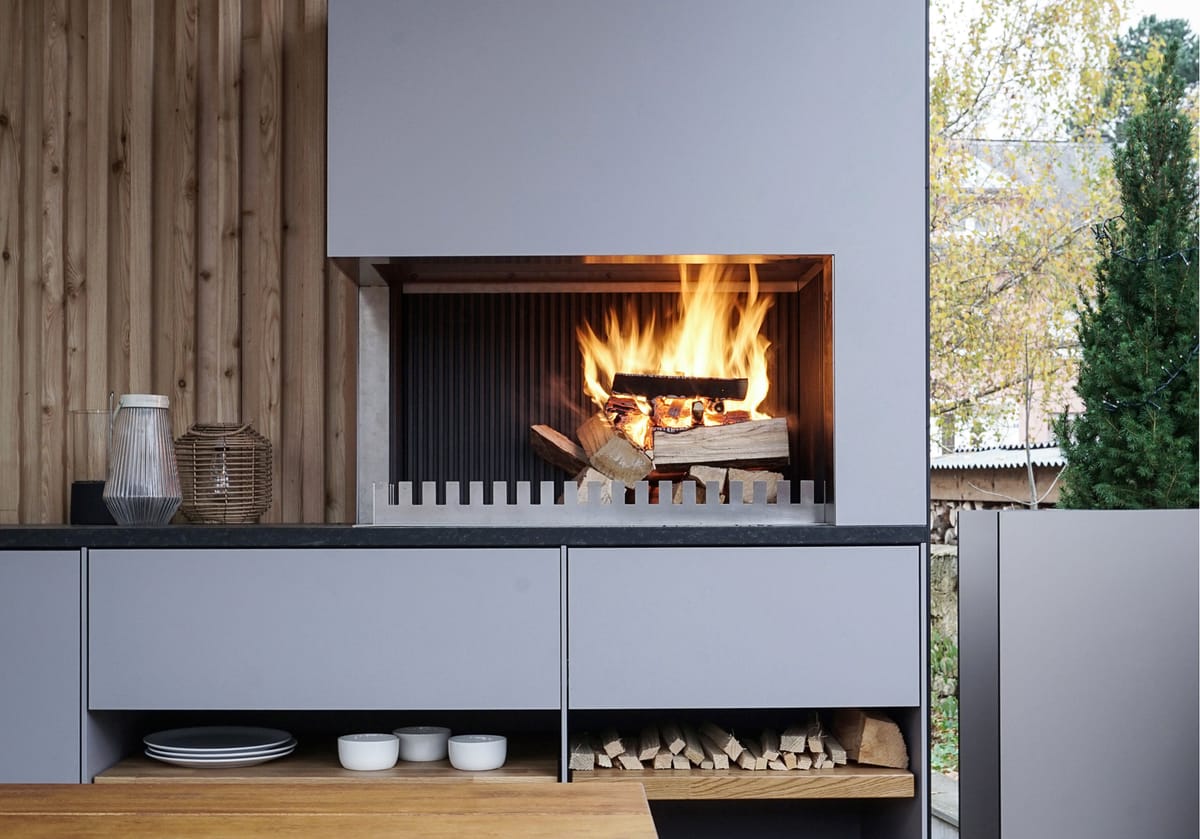 Wood fire in a light gray grill-and-fire element above open shelves with plates and firewood