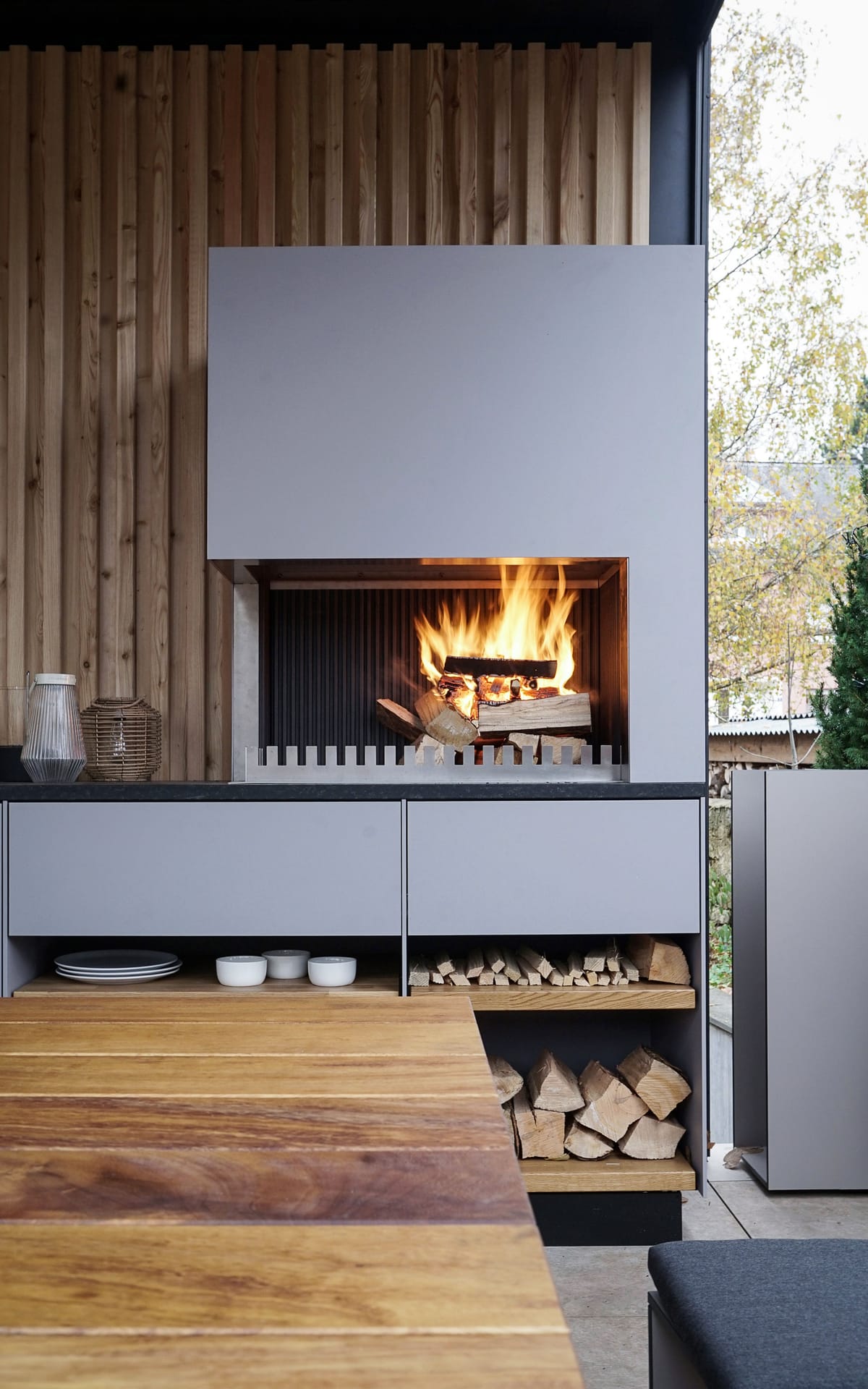 Wood fire in a light gray grill-and-fire element above open shelves with plates and firewood