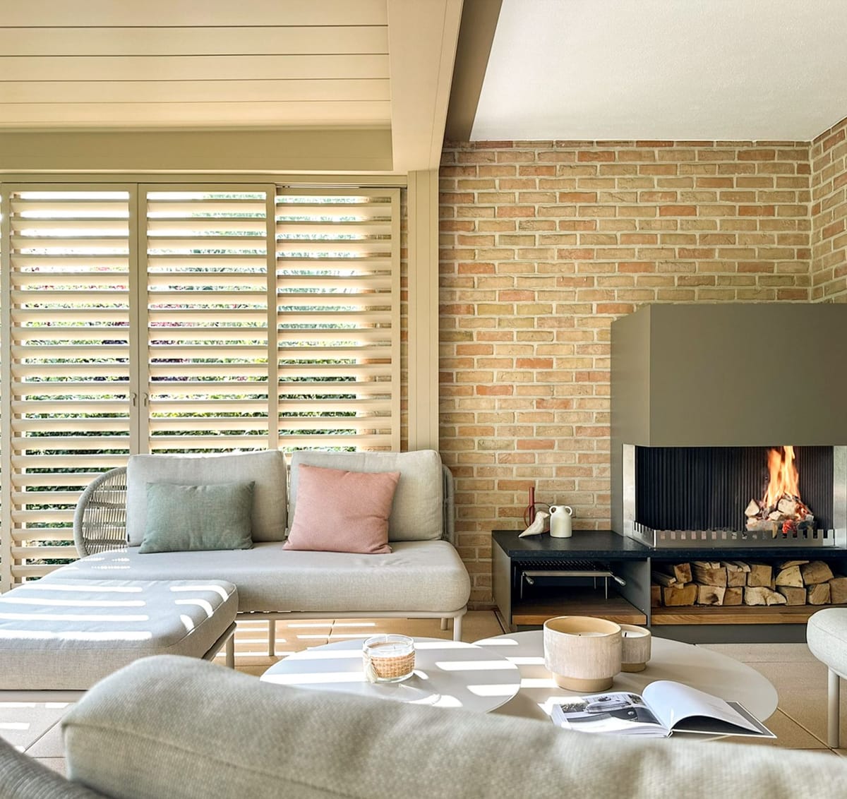 Covered lounge with light sofas, round tables and a wood fire burning in a gray steel fireplace unit with log storage