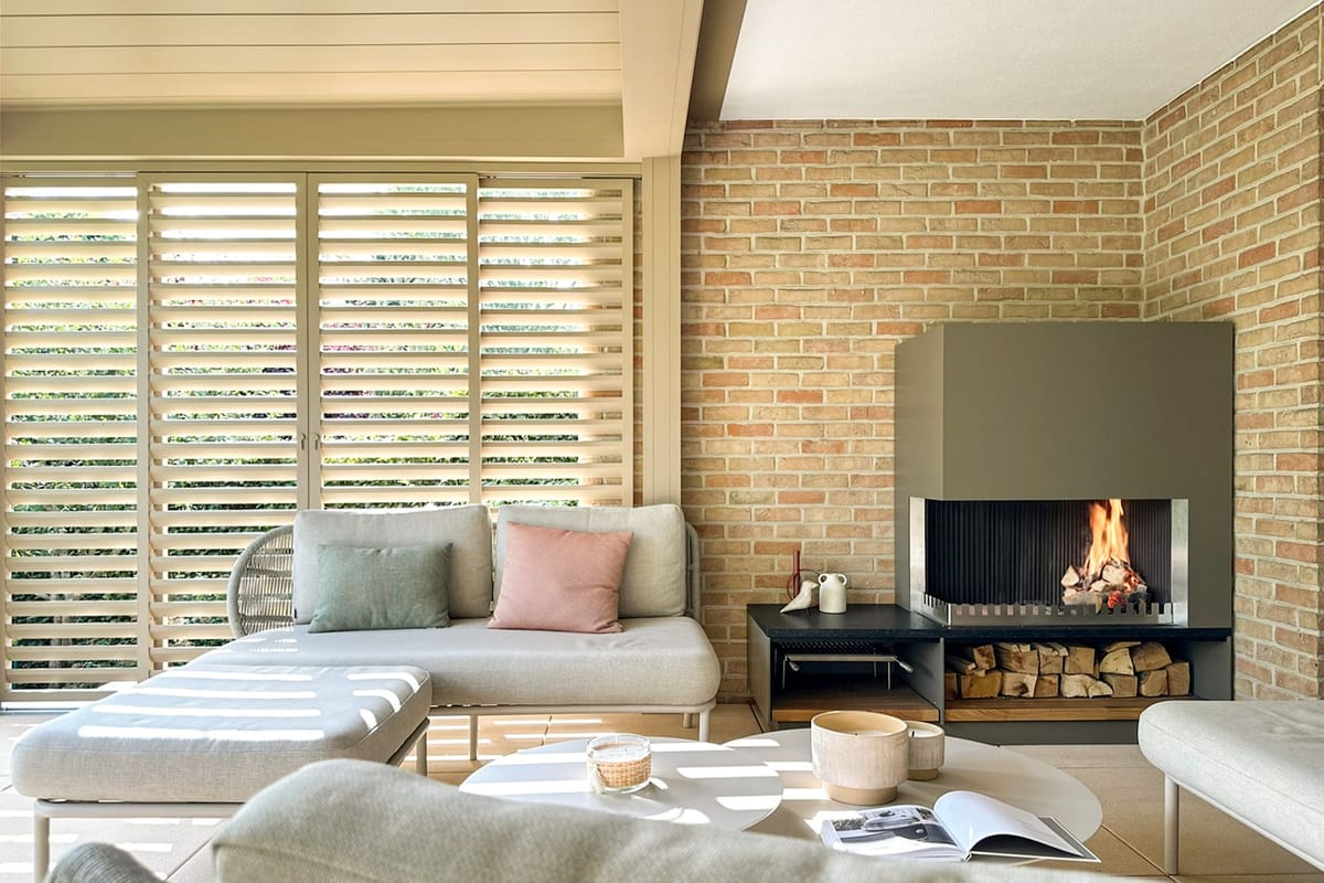 Covered lounge with light sofas, round tables and a wood fire burning in a gray steel fireplace unit with log storage