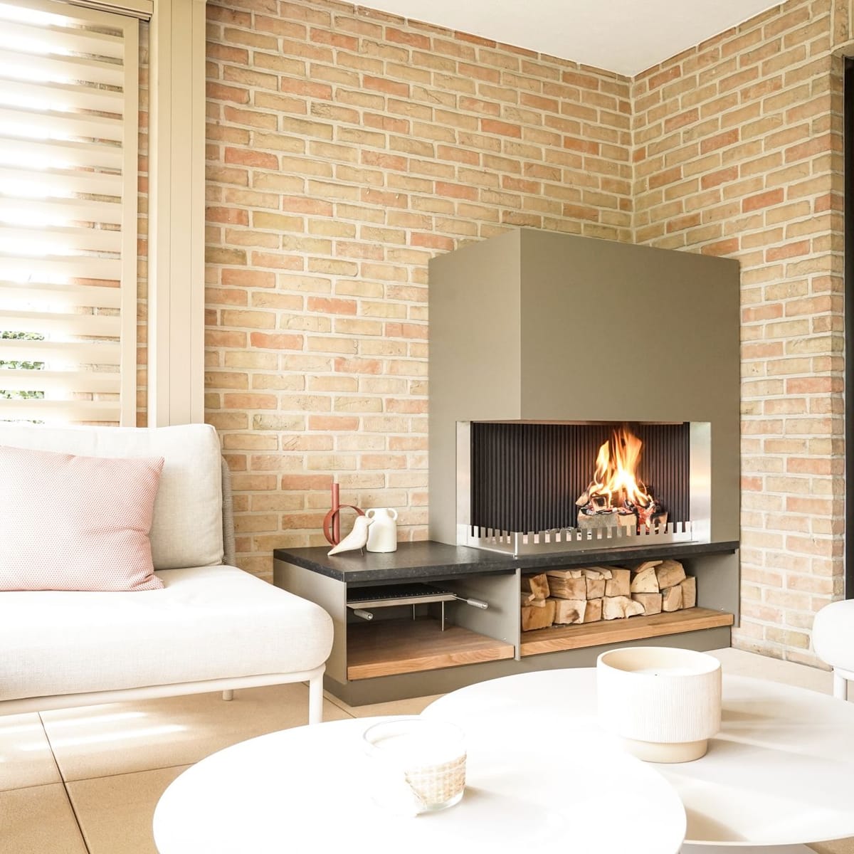 Wood fire in a khaki steel fireplace unit with a firewood shelf in a brick-walled lounge with white sofas