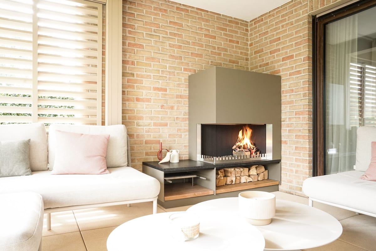 Wood fire in a khaki steel fireplace unit with a firewood shelf in a brick-walled lounge with white sofas