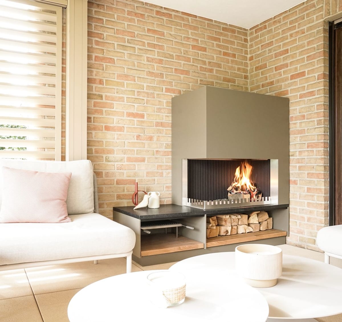 Wood fire in a khaki steel fireplace unit with a firewood shelf in a brick-walled lounge with white sofas